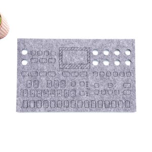 May include: A gray felt panel with cutouts and etched lines, possibly for a synthesizer or electronic device. The panel has various rectangular and circular openings. A small potted succulent is in the upper left corner.
