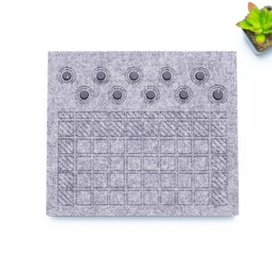 May include: A gray felt board with a grid pattern and nine black buttons. The board is designed for playing a game.