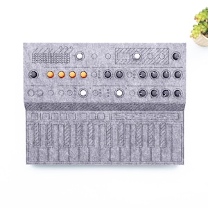 May include: A gray felt synthesizer with a keyboard, knobs, and buttons. The design includes orange lights and a detailed illustration of the instrument's components. A small succulent plant in a white pot is in the upper right corner.