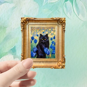 May include: A miniature gold picture frame with an elaborate design, displaying a painting of a black cat surrounded by blue tulips and yellow flowers. The frame is held in a hand.