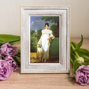 May include: A white framed picture with a painting of a woman in a white dress standing in a garden. The woman is holding a bouquet of flowers and is wearing a shawl. The painting is set against a backdrop of green trees and a blue sky.