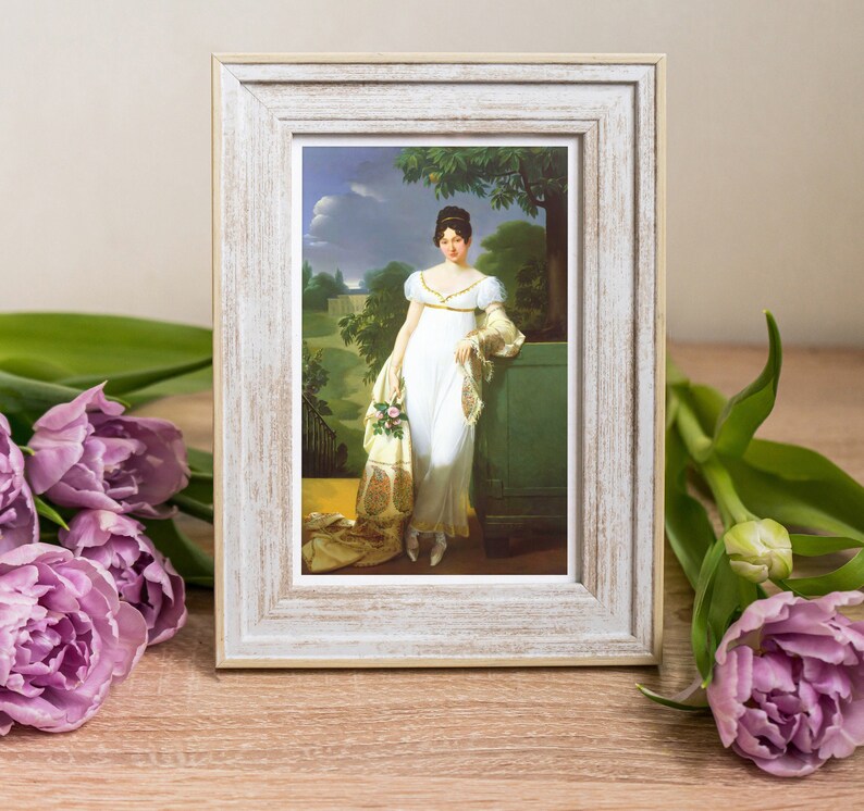 May include: A white framed picture with a painting of a woman in a white dress standing in a garden. The woman is holding a bouquet of flowers and is wearing a shawl. The painting is set against a backdrop of green trees and a blue sky.