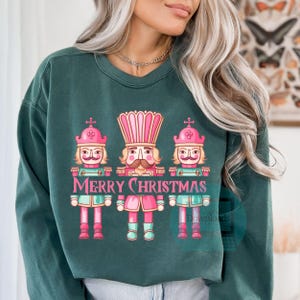Pode incluir: Uma camisa de frio verde com um design de Natal com três figuras de quebra-nozes com chapéus cor-de-rosa e o texto "Merry Christmas".