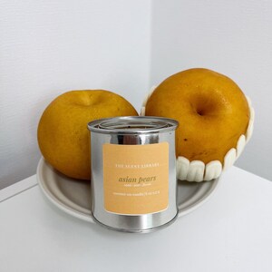 May include: A silver tin candle with a yellow label that reads "THE SCENT LIBRARY asian pears apple pear floral coconut soy candle | 8 oz | 227 g". Two yellow pears are in the background.