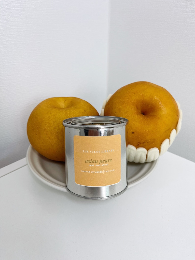 May include: A silver tin candle with a yellow label that reads "THE SCENT LIBRARY asian pears apple pear floral coconut soy candle | 8 oz | 227 g". Two yellow pears are in the background.