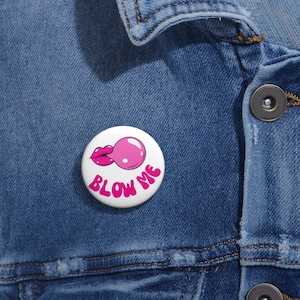 Può includere: Un bottone bianco con un disegno di labbra e una bolla di gomma da masticare rosa. Le parole "BLOW ME" sono scritte in rosa. Il bottone è appuntato su una giacca di jeans blu.