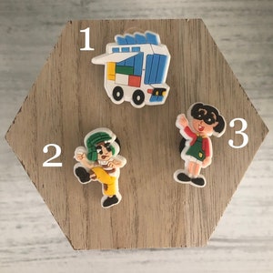 May include: Three colorful cartoon character shoe charms. The charms are numbered 1, 2, and 3. Charm 1 is a blue and white delivery truck. Charm 2 is a green and yellow cartoon character. Charm 3 is a red, yellow, and blue cartoon character.