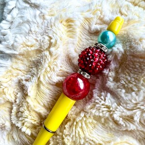 May include: A decorative pen with a yellow barrel and a beaded top. The beaded top features a large red bead, a cluster of red rhinestones, a teal bead, and a yellow bead. The pen is set against a fluffy, cream-colored background.