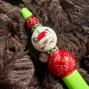 Può includere: Una penna verde con un design a tema natalizio. La penna presenta una perla bianca con un unicorno con un cappello da Babbo Natale, dettagli rossi e verdi e una perla rossa glitterata.