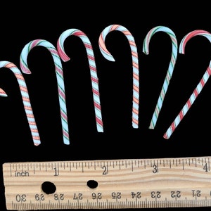 May include: Seven candy canes with red, white, and blue stripes. The candy canes are arranged in a semi-circle on a black background. A wooden ruler with inch markings is below the candy canes.