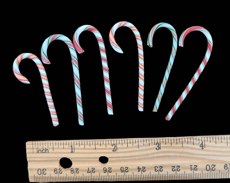 May include: Seven candy canes with red, white, and blue stripes. The candy canes are arranged in a semi-circle on a black background. A wooden ruler with inch markings is below the candy canes.
