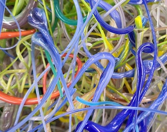 May include: A close-up of a tangled mass of colorful glass rods. The rods are in various shades of blue, green, yellow, and orange. The glass rods are arranged in a random pattern, creating a visually interesting and abstract design.