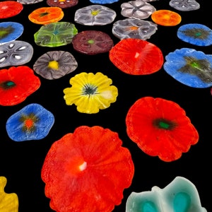 May include: Assortment of colorful glass flower-shaped coasters. The coasters are various shades of red, blue, green, yellow, and white. The coasters are arranged on a black background.