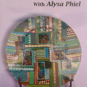 May include: A decorative plate with a colorful, geometric design. The plate features a patchwork of teal, green, brown, and white stripes and squares. The text "Mastering On Edge With Alysa Phiel" is at the top.