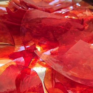 May include: A collection of translucent, jagged shards of red and orange glass. The pieces vary in size and shape, featuring smooth, reflective surfaces with sharp, irregular edges that catch the light, creating a layered, crystalline appearance.