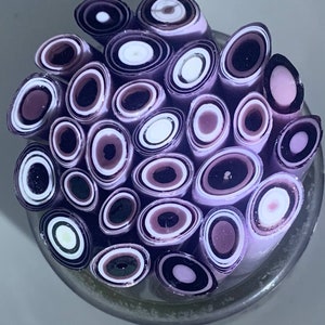 May include: A close-up of a bundle of purple, white, and black polymer clay canes. The canes are arranged in a circular pattern, showing the cross-section of each cane.