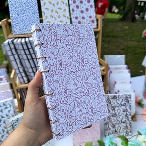 May include: A notebook with a white cover and a repeating pattern of purple squiggles, bound with a spiral. Other notebooks with different patterns, such as blue dots, yellow pineapples, and pink flowers, are visible in the background. The notebook is held by a person.