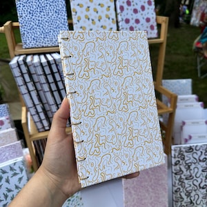 May include: A white notebook with a gold squiggly pattern is held up, showcasing a range of patterned notebooks. Other designs include blue dots, yellow pineapples, and pink floral patterns. The notebooks are bound with metal rings.