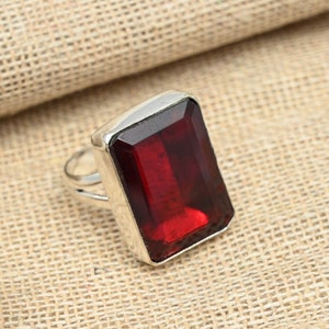 May include: A silver ring with a large, rectangular, deep red gemstone. The gemstone is set in a silver bezel. The ring has a simple design. It is displayed on a textured, tan surface.
