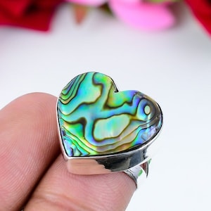 Puede incluir: Un anillo de plata con una incrustación de concha de abulón en forma de corazón. La concha tiene colores iridiscentes verdes, azules y dorados.