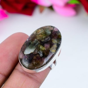 Puede incluir: Anillo ovalado con una gran piedra preciosa multicolor. La piedra presenta tonos de verde, morado y dorado, engastada en una banda de plata. El anillo se muestra sobre un fondo suave y borroso.