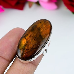 May include: A silver ring featuring a large, oval-shaped labradorite gemstone. The stone has a warm, brown and orange color with iridescent flashes. The ring's band is a simple, polished silver.