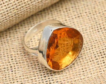 May include: A silver ring featuring a large, faceted, orange-yellow gemstone. The gemstone is set in a bezel setting, and the ring band is smooth and polished. The ring is displayed on a textured, tan surface.