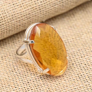 May include: A silver ring featuring a large, faceted, oval-shaped gemstone in shades of yellow and orange. The gemstone is set in a silver band, creating a striking contrast. The ring is displayed on a textured, tan surface.