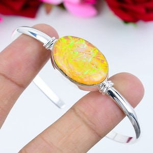 May include: A silver bangle bracelet with a large oval-shaped opal gemstone. The opal is a vibrant orange color with green and yellow flashes.