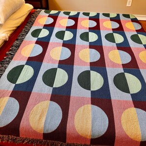 May include: A colorful woven blanket with a geometric pattern of half-circles in various colors, including pink, blue, green, and yellow. The blanket has a fringed edge and is displayed on a bed. The design is a grid of squares and circles.