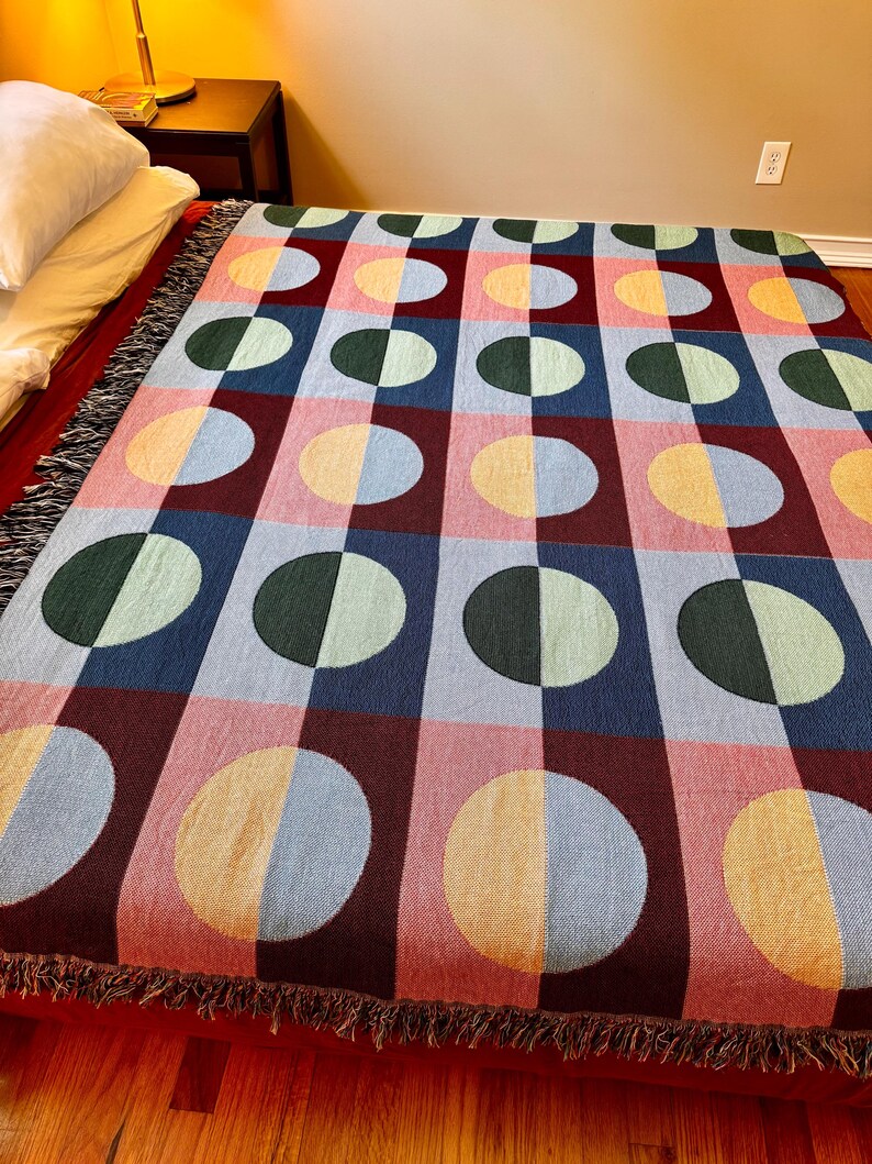 May include: A colorful woven blanket with a geometric pattern of half-circles in various colors, including pink, blue, green, and yellow. The blanket has a fringed edge and is displayed on a bed. The design is a grid of squares and circles.