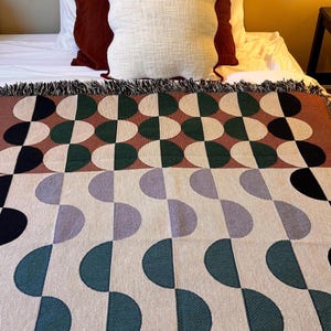 May include: A woven throw blanket with a geometric pattern of green, brown, purple, and black half circles on a cream background. The blanket has a fringe border.