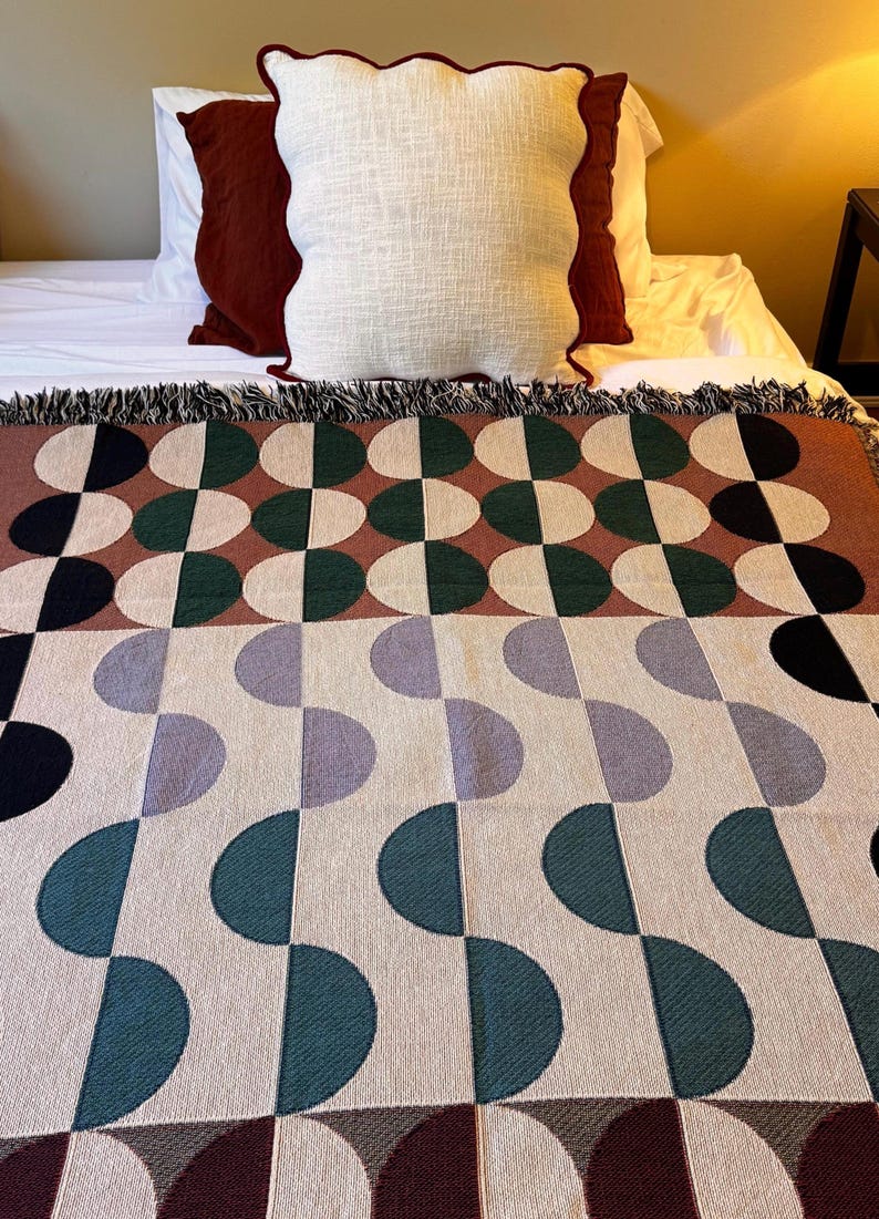 May include: A woven throw blanket with a geometric pattern of green, brown, purple, and black half circles on a cream background. The blanket has a fringe border.
