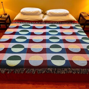 May include: A colorful woven blanket with a geometric pattern of half-circles in various colors, including blue, green, pink, and yellow. The blanket has a fringed edge and is displayed on a bed with white pillows.