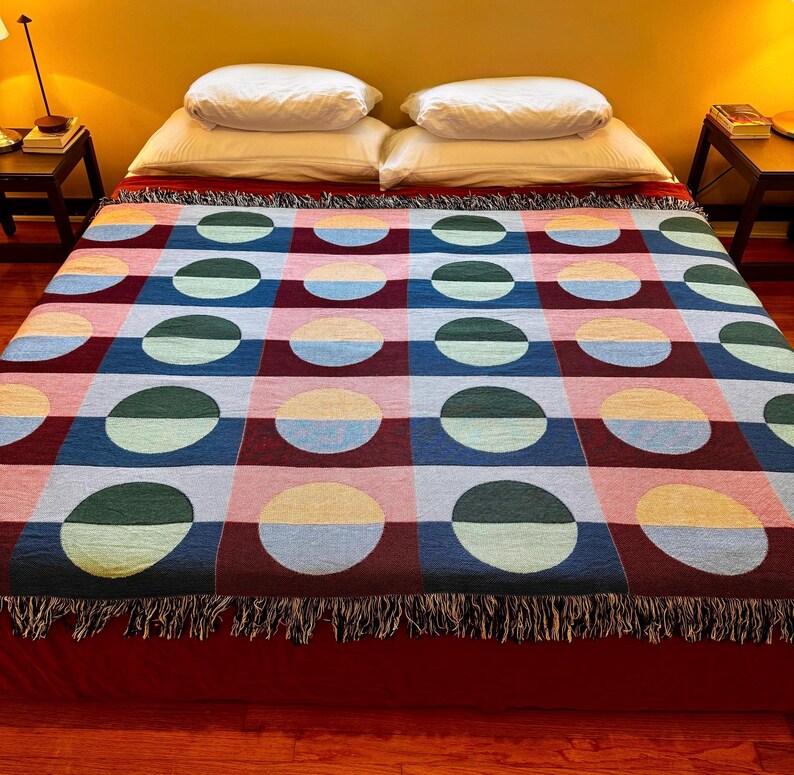 May include: A colorful woven blanket with a geometric pattern of half-circles in various colors, including blue, green, pink, and yellow. The blanket has a fringed edge and is displayed on a bed with white pillows.