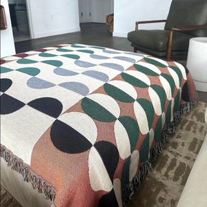 May include: A woven throw blanket with a geometric pattern of green, blue, black, white, and orange semi-circles. The blanket has a fringe border.