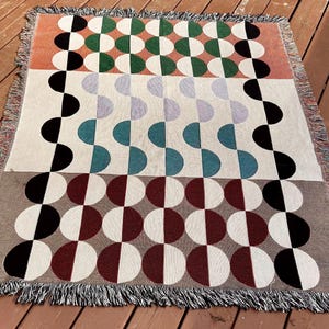 May include: A woven throw blanket with a geometric pattern of black, white, red, green, and blue semi-circles. The blanket has a fringe border.