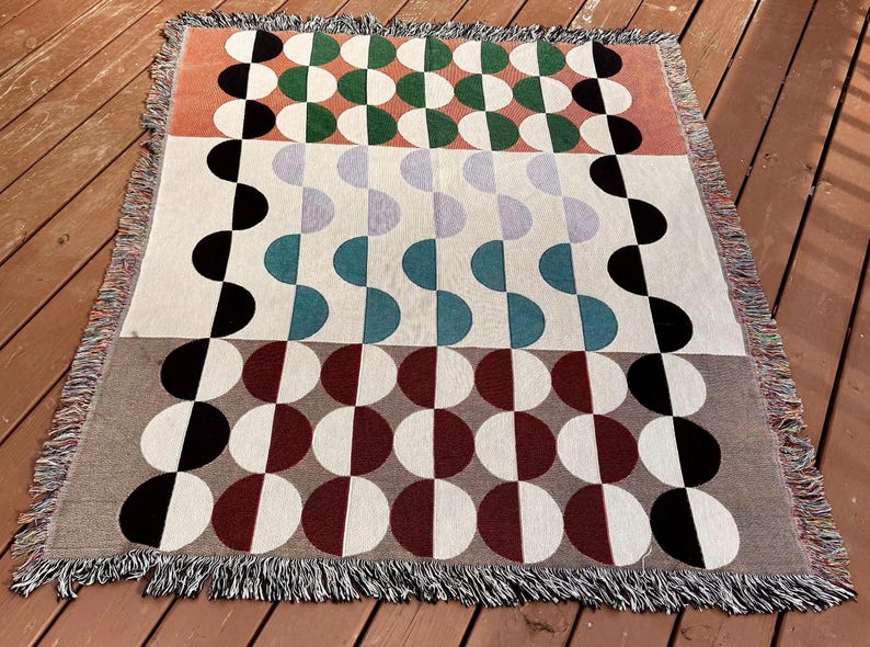 May include: A woven throw blanket with a geometric pattern of black, white, red, green, and blue semi-circles. The blanket has a fringe border.