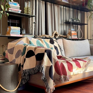 May include: A colorful geometric throw blanket with fringe edges draped over a light gray couch. The blanket features a pattern of red, black, white, and green circles and semi-circles.