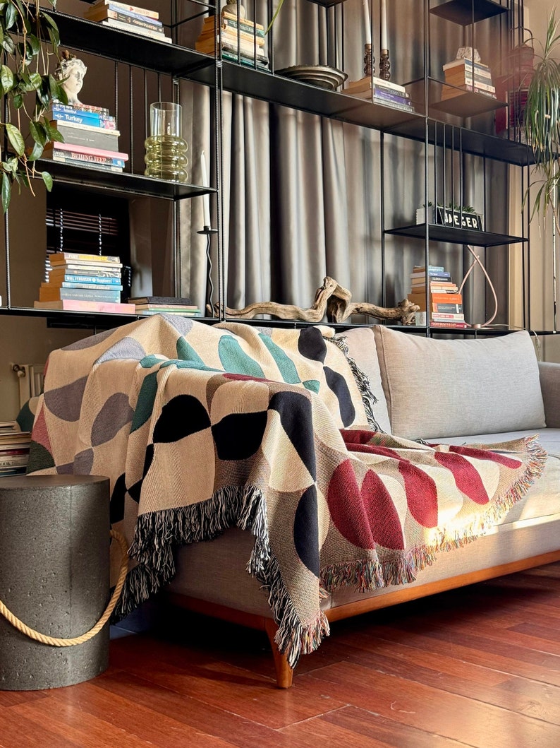 May include: A colorful geometric throw blanket with fringe edges draped over a light gray couch. The blanket features a pattern of red, black, white, and green circles and semi-circles.