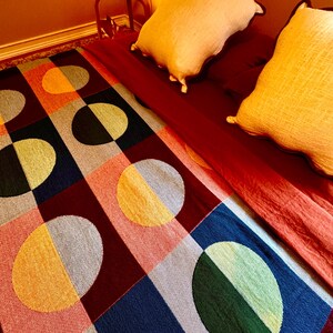 May include: A colorful geometric patterned rug with large circles and squares in shades of red, blue, green, yellow, and gray. Two cream-colored pillows rest on a red bedspread. A small red side table with a lamp is in the background.