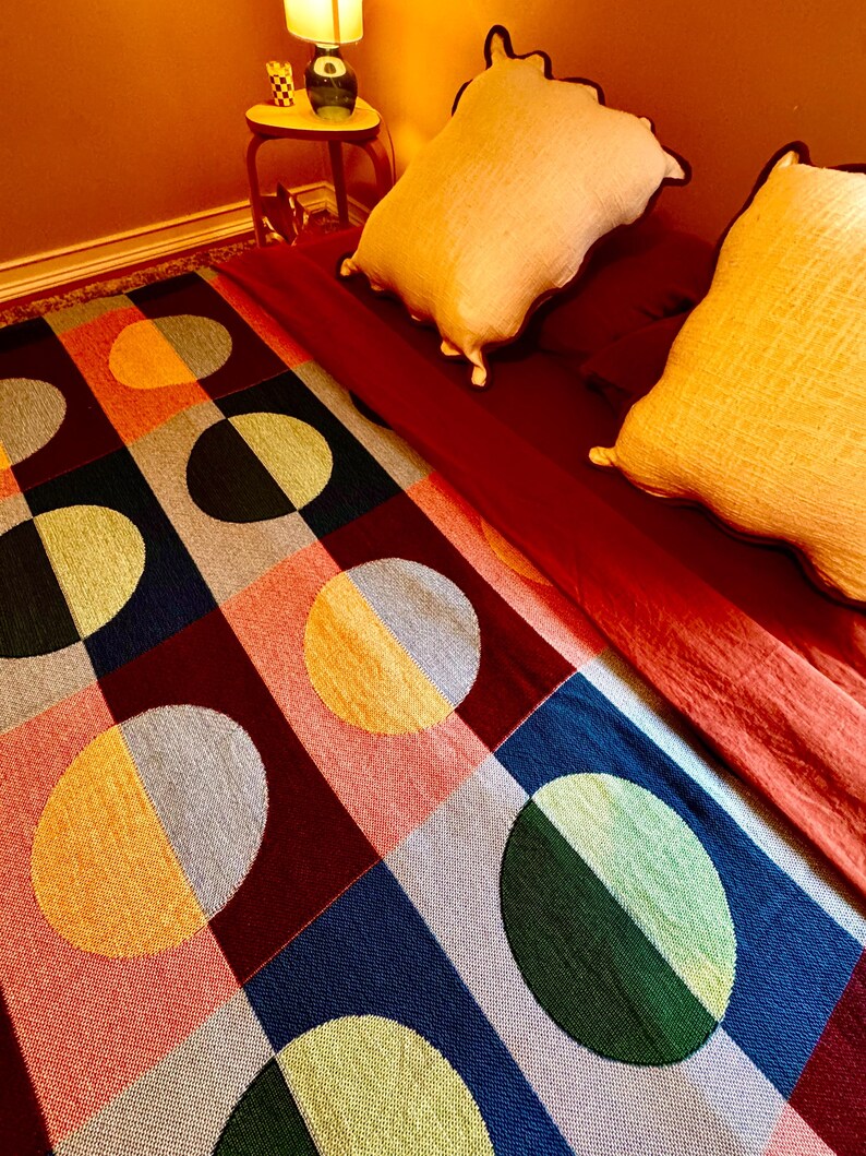 May include: A colorful geometric patterned rug with large circles and squares in shades of red, blue, green, yellow, and gray. Two cream-colored pillows rest on a red bedspread. A small red side table with a lamp is in the background.