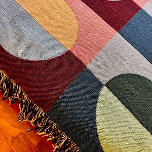 May include: A woven throw blanket with a geometric pattern of circles and squares in shades of burgundy, pink, blue, green, and gold. The blanket has a fringe edge. The blanket is on a wooden surface.