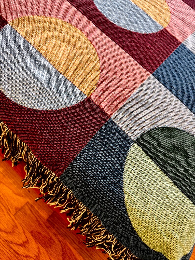May include: A woven throw blanket with a geometric pattern of circles and squares in shades of burgundy, pink, blue, green, and gold. The blanket has a fringe edge. The blanket is on a wooden surface.