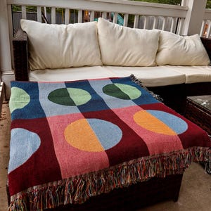 May include: A colorful woven throw blanket with a geometric pattern of circles and squares in shades of blue, red, pink, green, and orange. The blanket has a fringed edge and is draped over a dark brown ottoman.