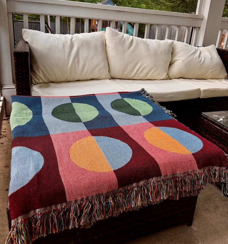 May include: A colorful woven throw blanket with a geometric pattern of circles and squares in shades of blue, red, pink, green, and orange. The blanket has a fringed edge and is draped over a dark brown ottoman.