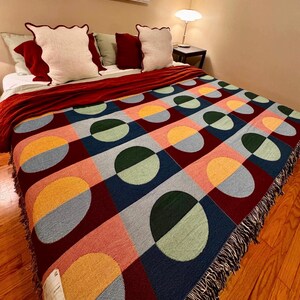 May include: A colorful woven blanket with a geometric pattern of circles and squares in shades of red, blue, green, yellow, and pink. The blanket has a fringed edge and is draped over a bed with pillows.