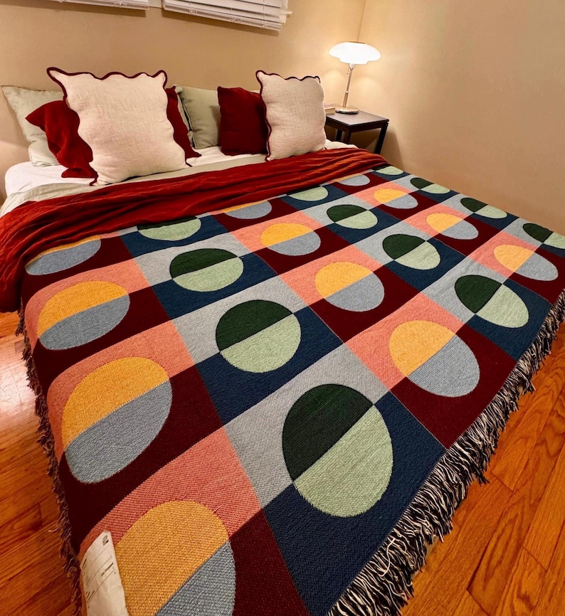 May include: A colorful woven blanket with a geometric pattern of circles and squares in shades of red, blue, green, yellow, and pink. The blanket has a fringed edge and is draped over a bed with pillows.