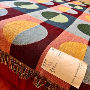 May include: A woven blanket with a colorful geometric pattern of squares and ovals in shades of red, blue, yellow, and green. The blanket has a fringed edge and measures 60x80 inches.