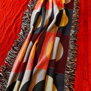 May include: A colorful throw blanket with a geometric pattern of circles and rectangles in shades of orange, yellow, gray, green, and navy blue. The blanket has a fringed edge and is draped over a red surface.